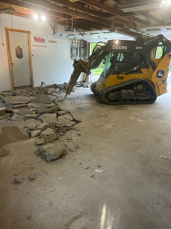 John Deere skid steer breaking up a concrete floor inside a garage during interior demolition..