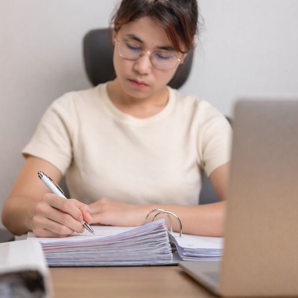 Woman writing notes with a stack of papers and laptop in front of her.