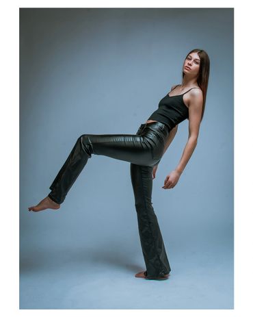 Model in black leather pants and tank top posing with one leg raised against a blue backdrop.