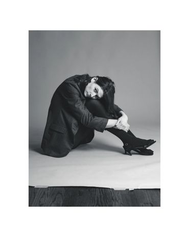 Black and white photo of a woman in a blazer sitting curled up on the floor.