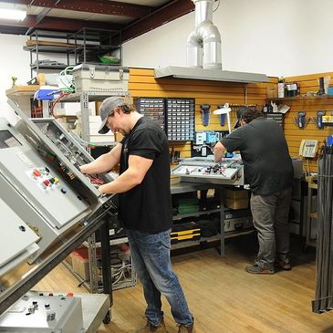 Two men working on electrical control panels in a workshop.