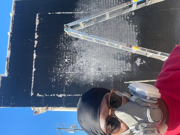 Person wearing a mask and sunglasses in front of a partially painted black brick wall with a ladder.