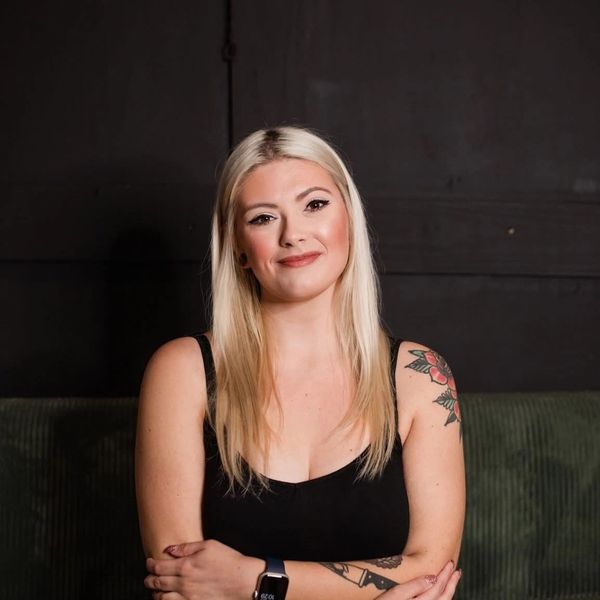 Blonde woman with tattoos smiling, wearing a black tank top against a dark background.