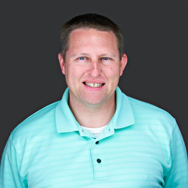 Smiling man in a light blue polo shirt against a dark background.