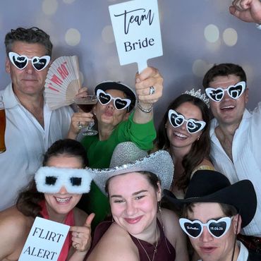 Group of friends at a fun photo booth wearing heart-shaped sunglasses and holding playful signs.