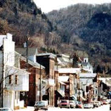 Small town street with parked cars and hills in the background.