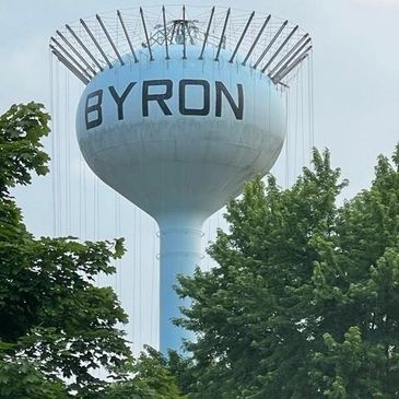 Water tower labeled BYRON surrounded by trees under a cloudy sky.