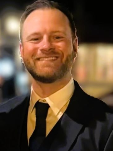Smiling man in formal attire with a blurred nighttime background.