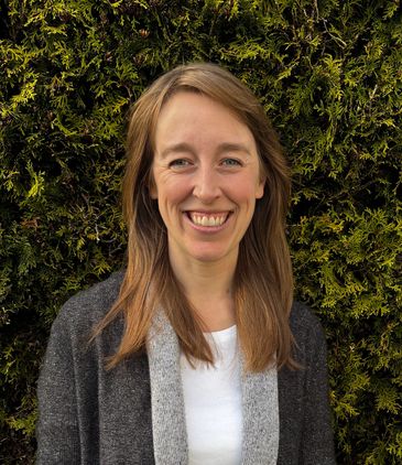 Smiling woman with long hair standing in front of a green leafy background.