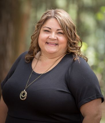 Smiling woman with curly hair wearing a black shirt and pendant necklace outdoors.
