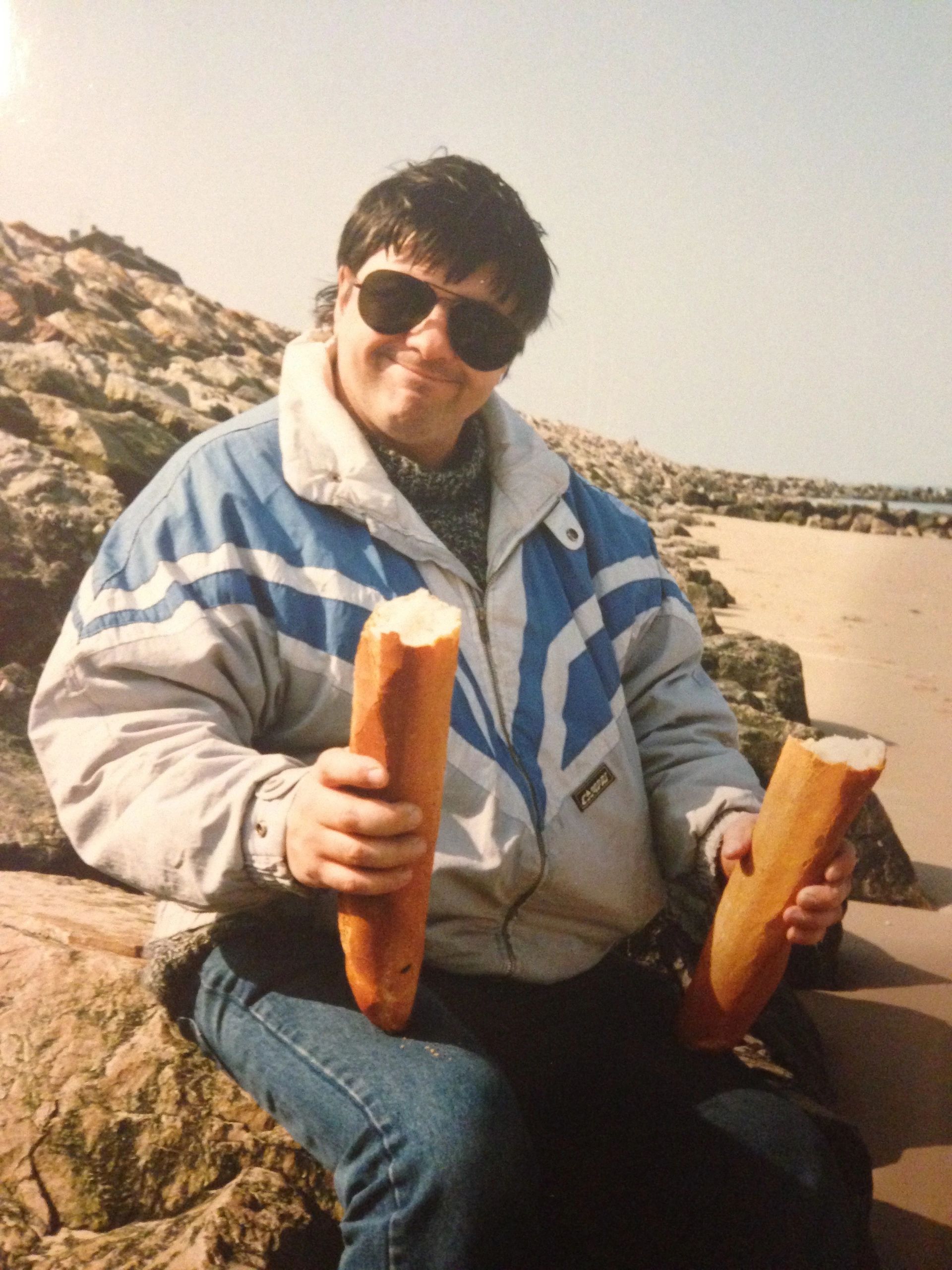 Man sitting on a rocky beach holding two baguettes, wearing sunglasses and a winter jacket.
