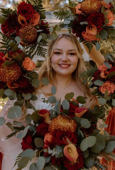 Bride framed by fall bridesmaid bouquets. red roses, Calla Lilies, Cremones, ferns, eucalyptus