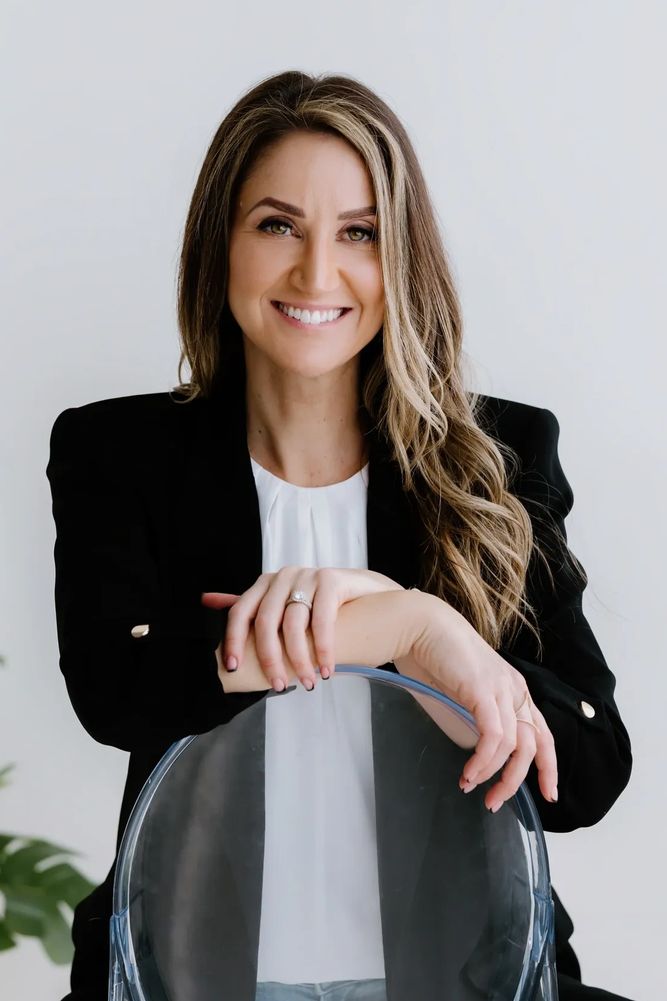 Smiling woman in black blazer poses with clear chair against white wall.