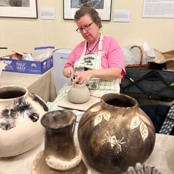 Lisa Lindler sculpts clay while seated behind a table of ceramics.
