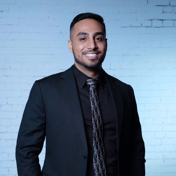 Man in black suit smiling against a blue brick wall.
