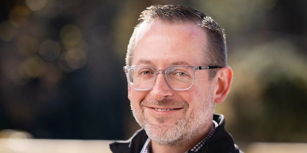 Smiling man with glasses outdoors in soft sunlight.