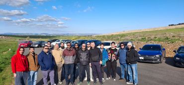 Group of people posing outdoors with cars and blue sky in the background.