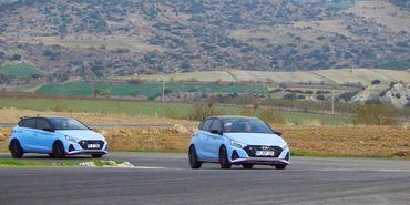 Two light blue compact cars racing on a track in a rural area.