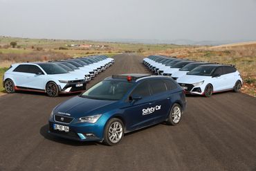 A blue safety car leads a lineup of white hatchback cars on a race track.