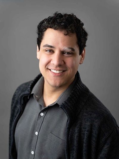 Smiling man in a dark cardigan and gray shirt against a gray background.