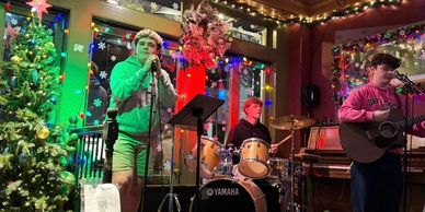 A festive band performs holiday music in a decorated room with Christmas lights and a tree.