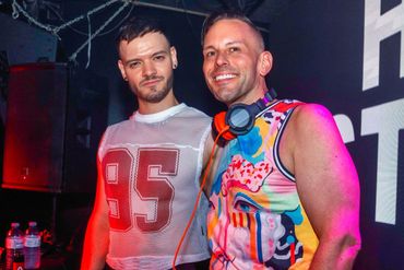 Two men smiling at a party, one wearing a mesh shirt and the other colorful clothes with headphones.