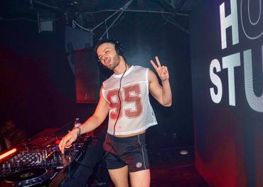 DJ in a mesh crop top and shorts posing with a peace sign at a club.