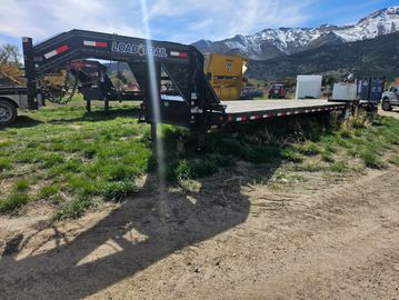 A flatbed Load Trail trailer parked on grassy ground with mountains in the background.