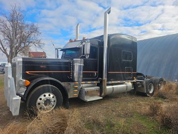Black semi-truck parked on grassy land with blue sky and clouds.