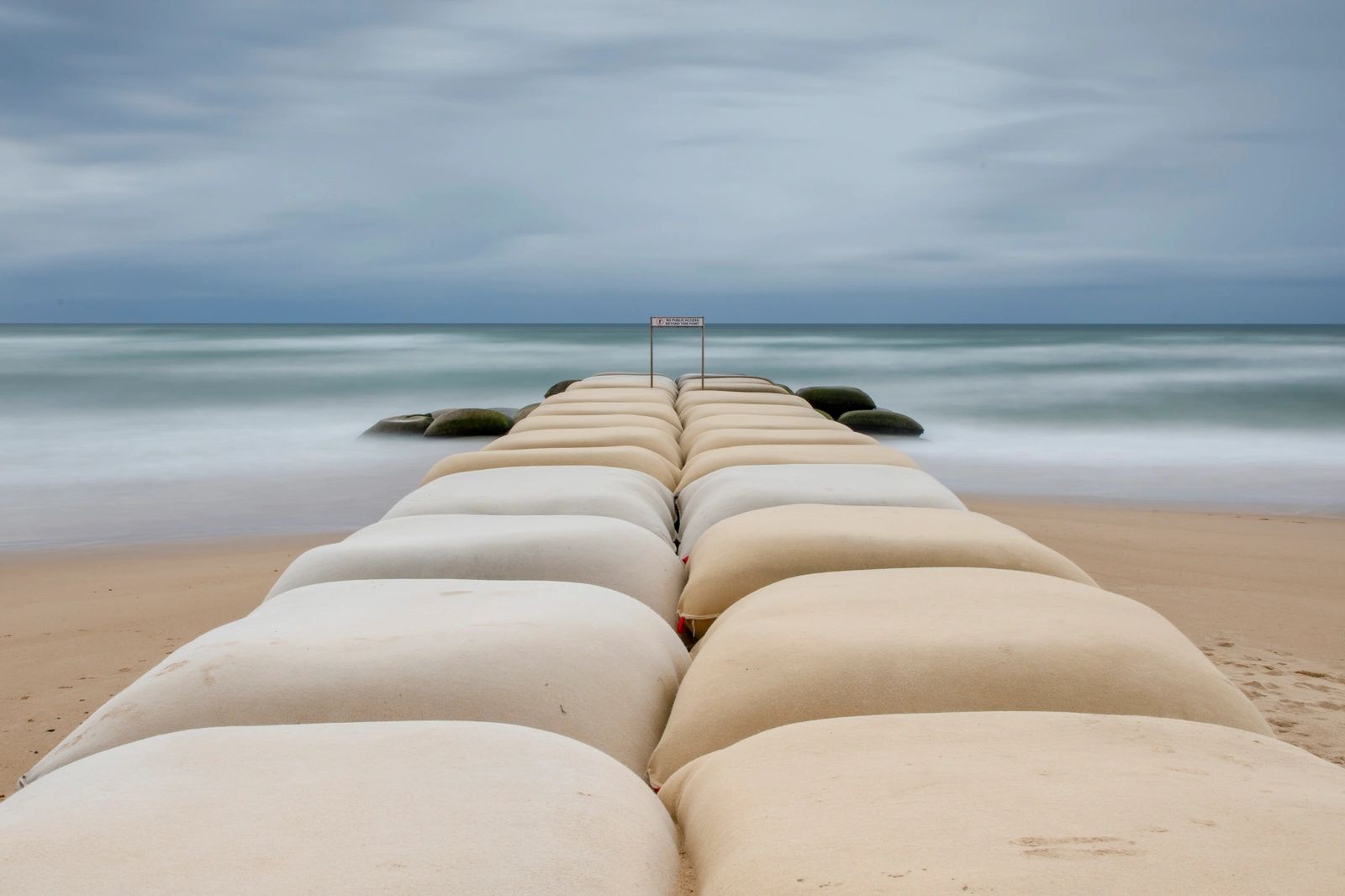 Long-exposure seascape with sandbag pathway leading into the ocean under a dramatic cloudy sky.
