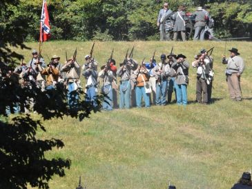 Reenactment of a historical battle with soldiers in period uniforms firing rifles.