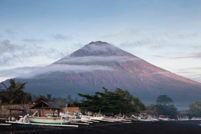 A towering volcano with a ring of clouds and boats on the shore below.