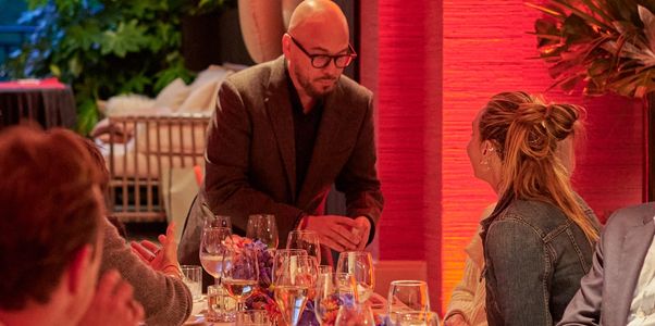 A man in glasses speaking to a woman at a warmly lit dinner table.