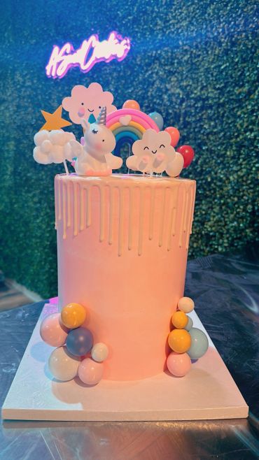 Tall pink drip cake with unicorn, rainbow, clouds, and colorful balls.