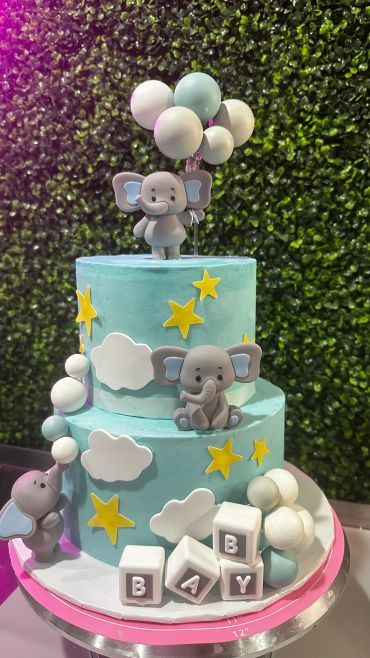 Two-tier blue baby shower cake with elephants and balloons.