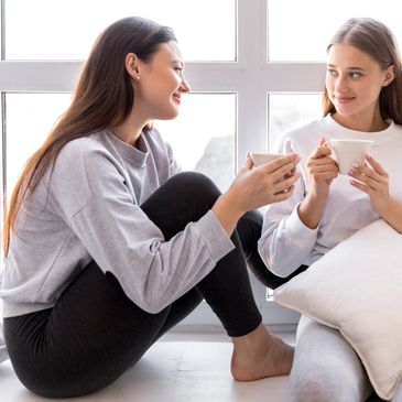 Two women in casual wear enjoying coffee and conversation by a bright window.
