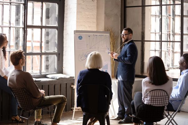 A medium shot shows a group of six adults in a meeting or training session in a well-lit room.