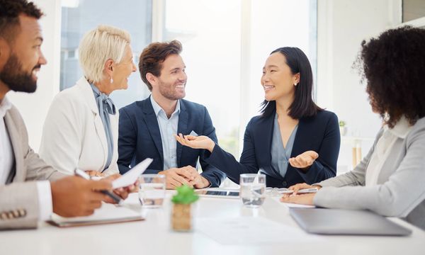 A medium shot shows a diverse group of five adults sitting around a white table in a bright office.