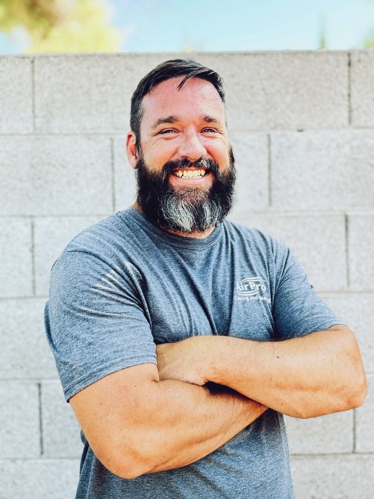 Smiling man with a beard in a gray t-shirt standing against a concrete wall.