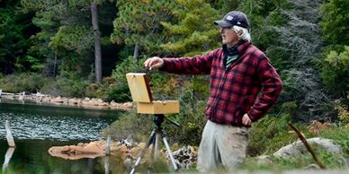 a painter in a red and black checked shirt stsanding at his easel and wearing a bal cap. Int eh back