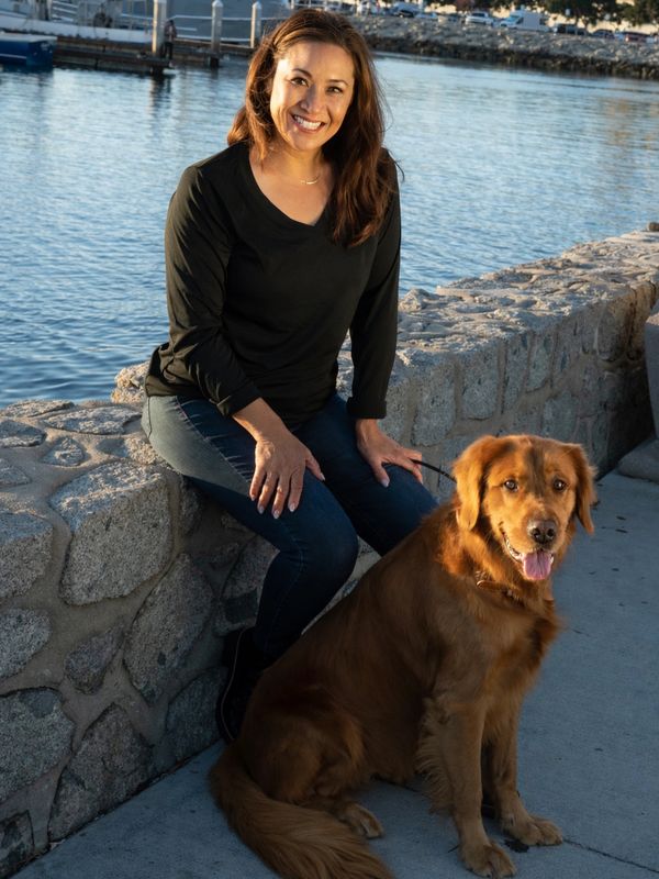 A woman smiles sitting on a stone wall by the water with a golden retriever.