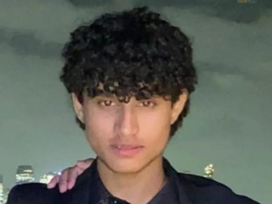 A young man with curly hair posing at night with a city skyline in the background.