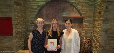 Three women posing indoors holding a cartoon picture.