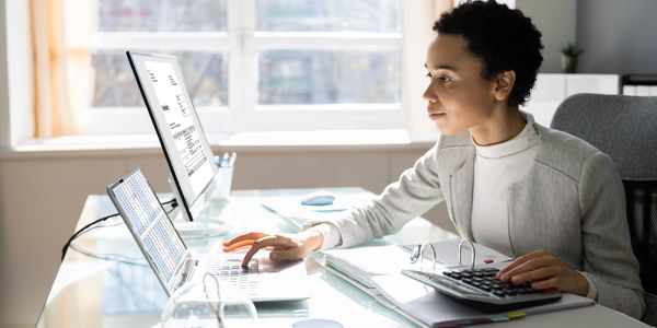 A woman sits at a modern desk, using a laptop and calculating with a keyboard