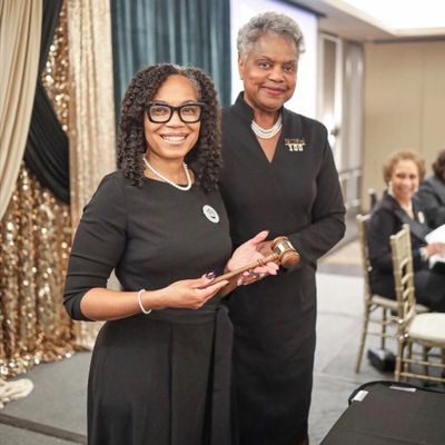 Two women in black dresses smiling, one holding a gavel at an event.
