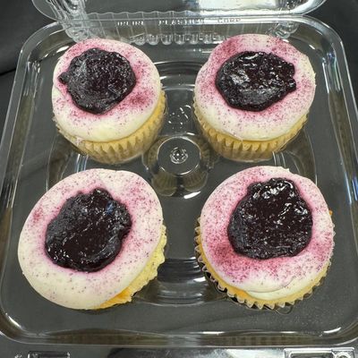 Four cupcakes with white frosting and dark berry topping in a plastic container.