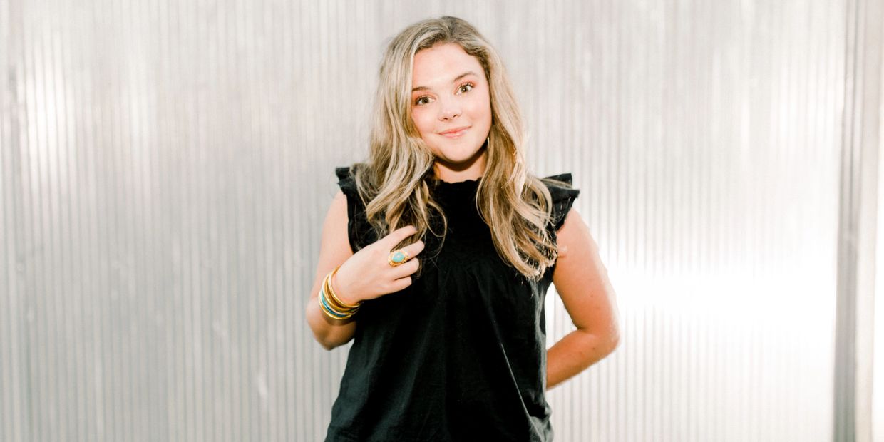 Young woman with blonde hair smiling in a black sleeveless top against a textured white background.