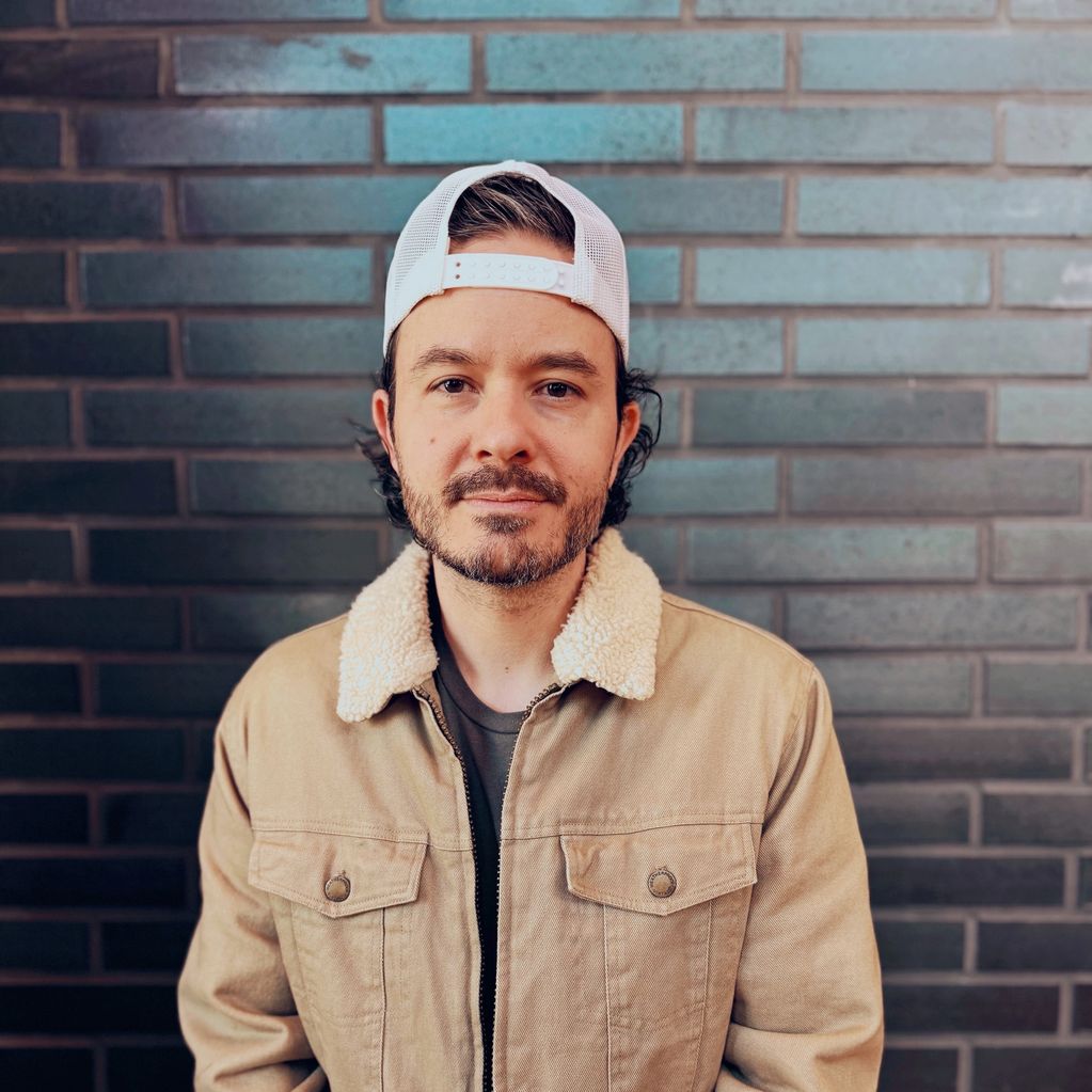 Man in a beige jacket and white cap posing against a brick wall.