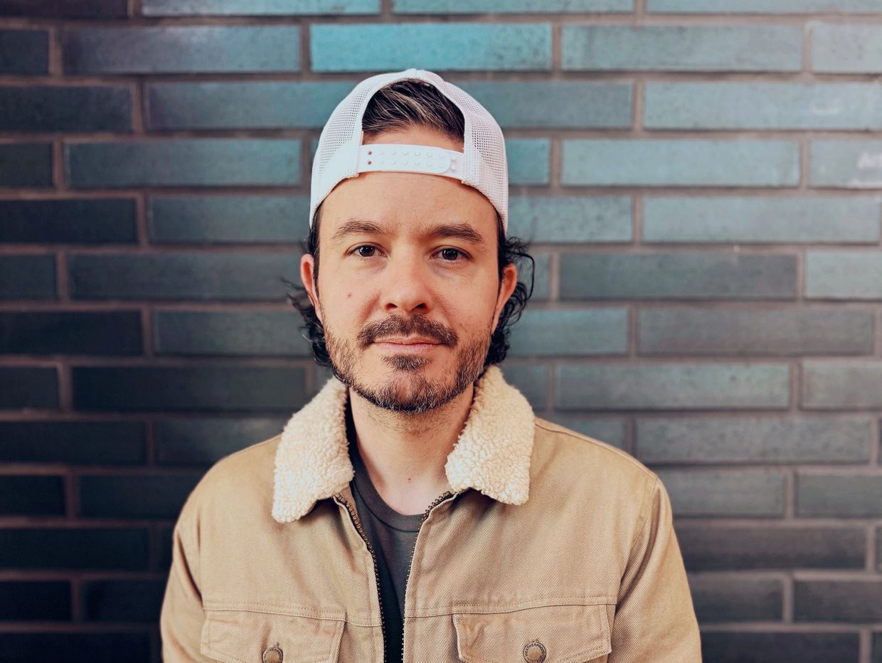 Man with a beard wearing a tan jacket and white cap against a brick wall.