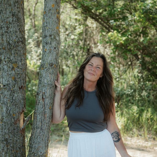 Woman in a gray top and white skirt stands beside a tree in a sunlit forest.